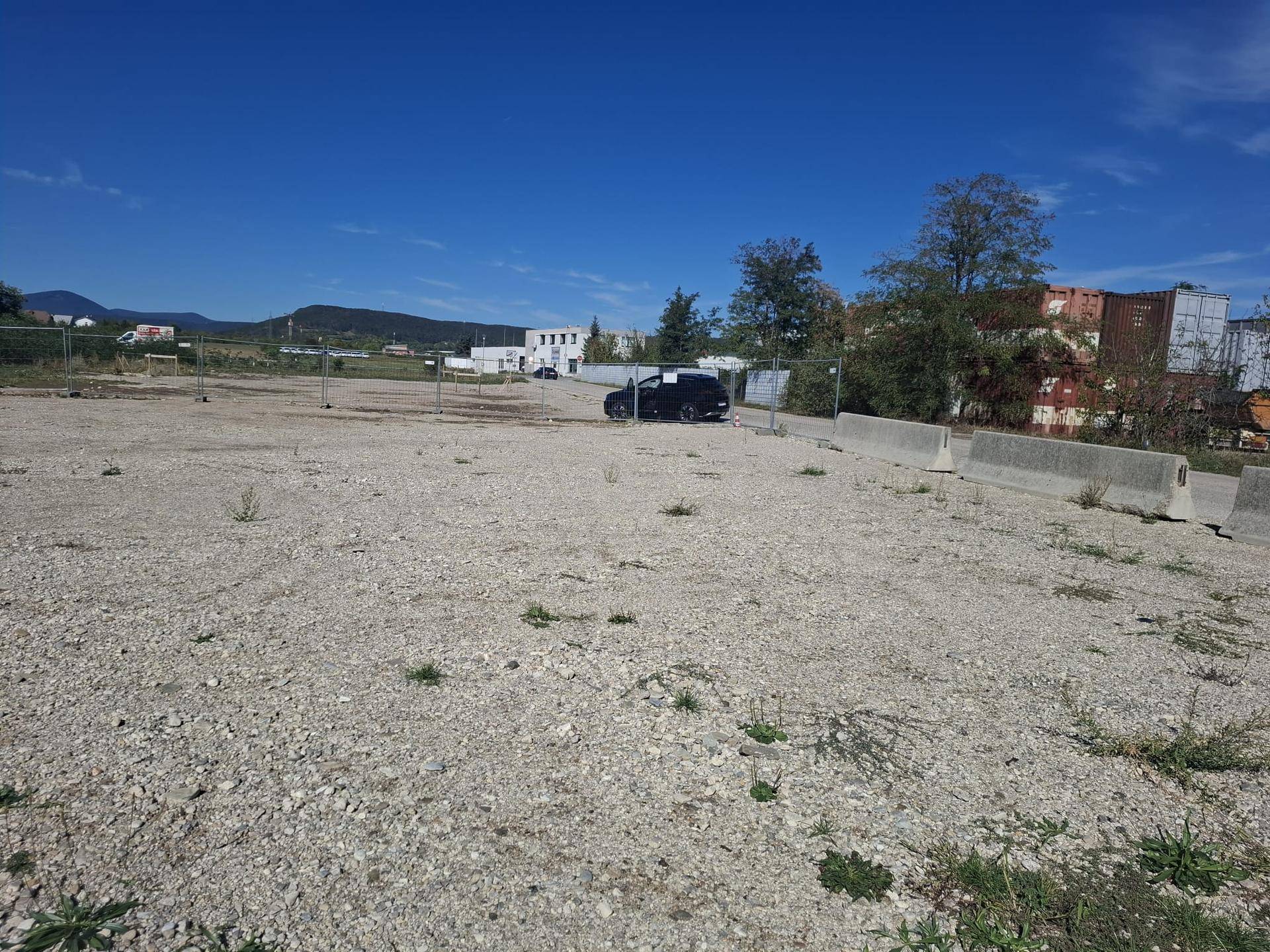 Großes, leeres Schottergrundstück mit Betonbarrieren, einem Auto und Industriegebäuden im Hintergrund.