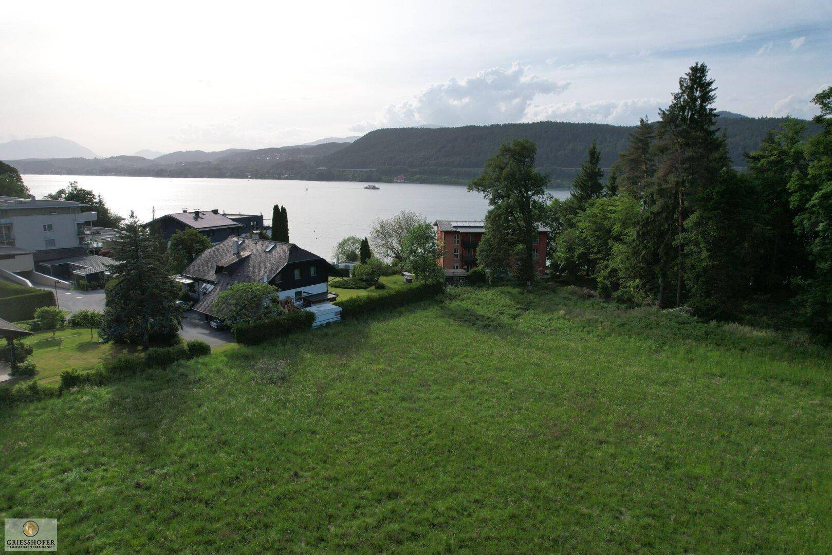 Luftaufnahme eines großen Grundstücks in Seenähe mit umliegenden Häusern und Bergen im Hintergrund.