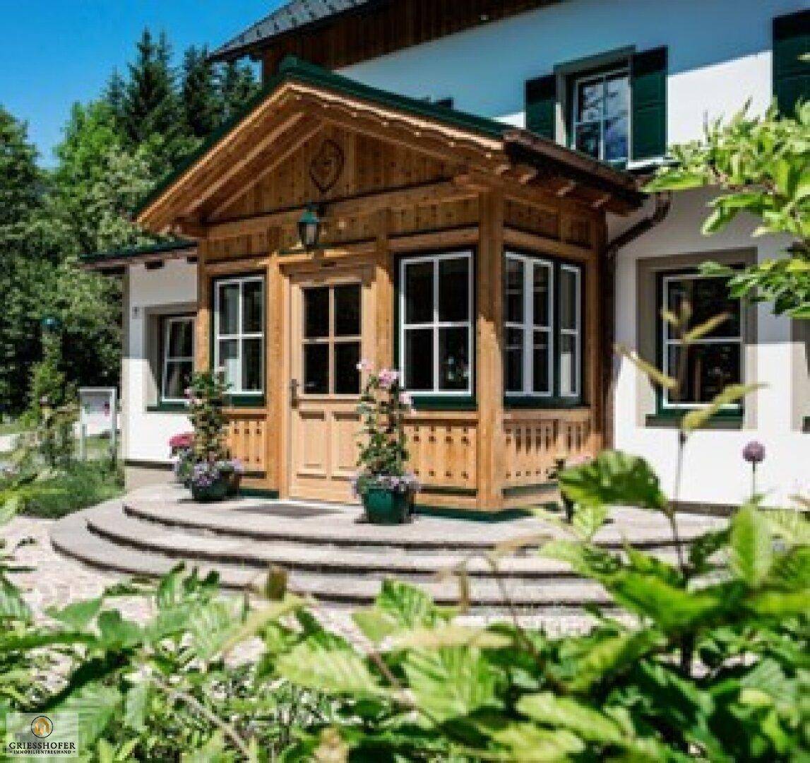 Traditionelles Haus mit Holzeingangsbereich und grüner Umgebung an einem sonnigen Tag.