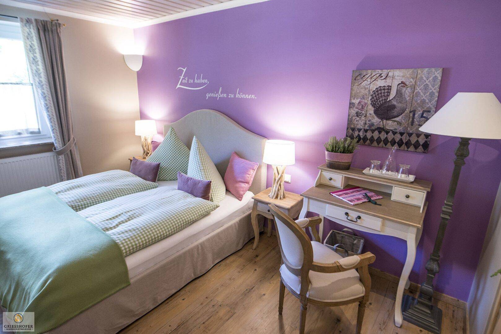 Gemütliches Schlafzimmer mit lila Akzentwand, Doppelbett und Holzboden.