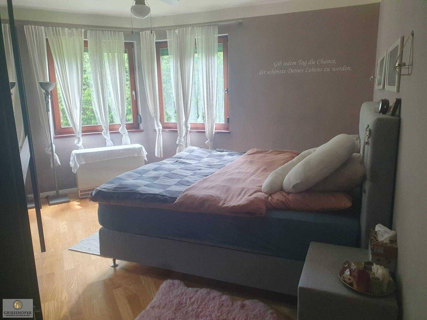 Helles Schlafzimmer mit großem Doppelbett, Holzboden und mehreren Fenstern mit Blick ins Grüne.