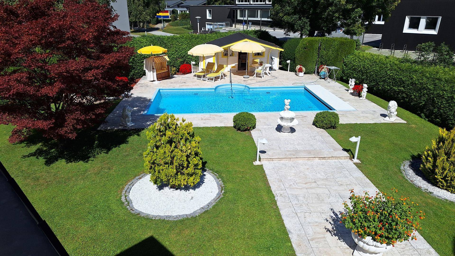 Gepflegter Gartenbereich mit großem Pool, Sonnenliegen, Sonnenschirmen und einem kleinen Poolhaus. Ein roter Baum und grüne Hecken umrahmen die Szene.