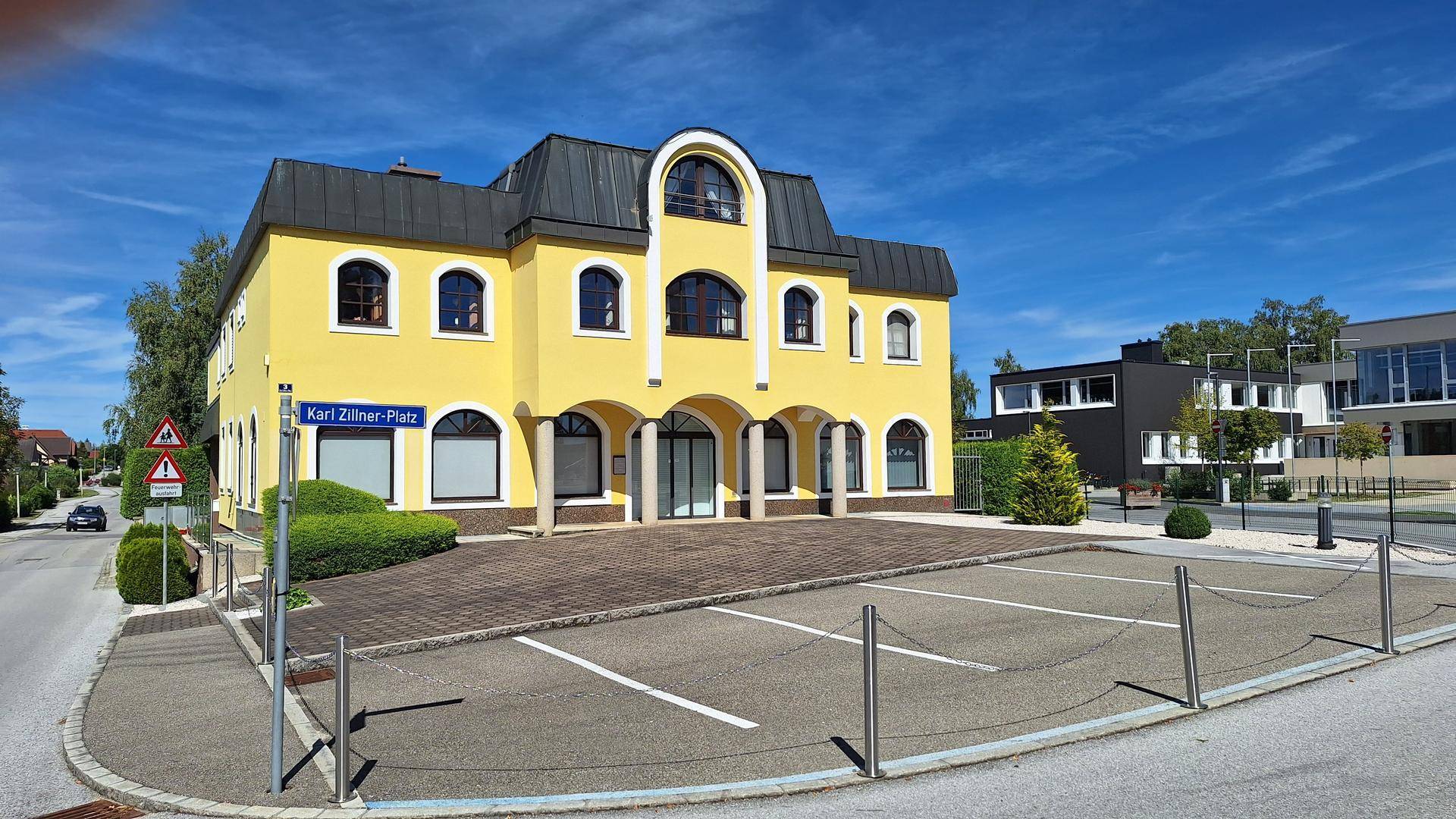 Gelbes Gebäude an einer Straßenecke mit Parkplätzen und einem Straßenschild unter blauem Himmel.