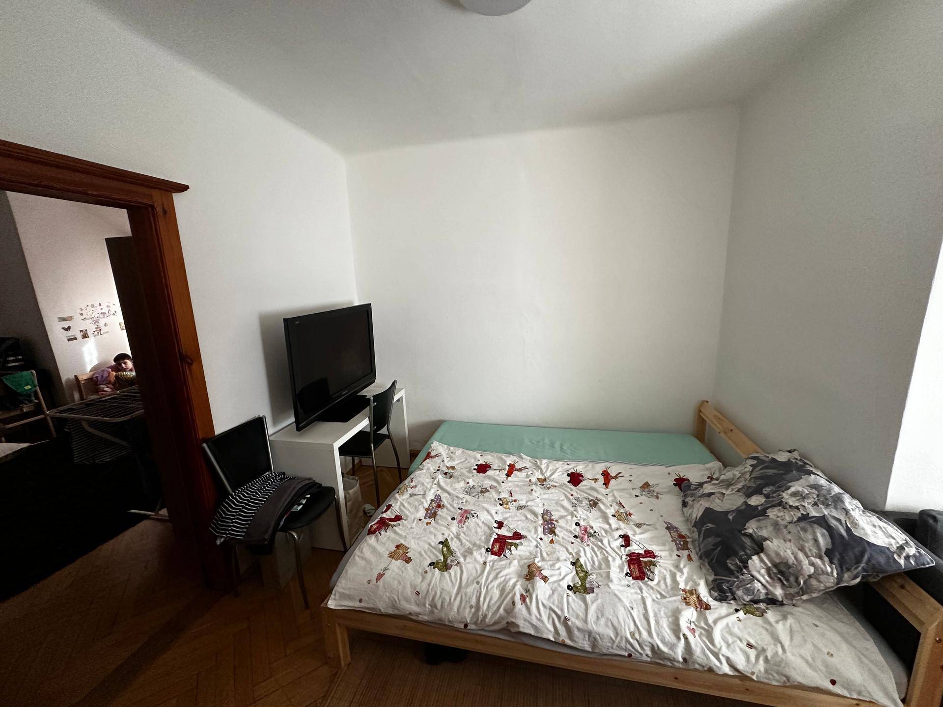 Helles Schlafzimmer mit Holzbett, Fernseher und Parkettboden, durch eine Tür mit einem weiteren Raum verbunden.