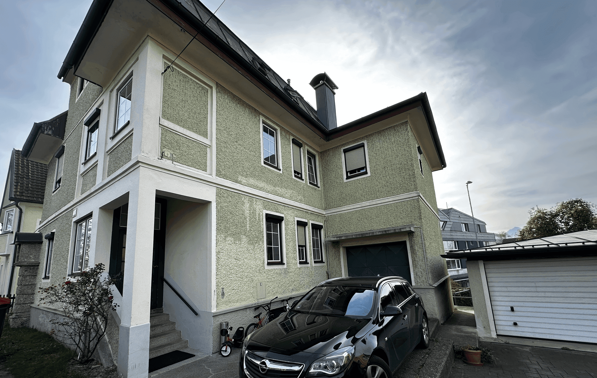 Grünes Wohnhaus mit Garage und gepflastertem Vorplatz unter bewölktem Himmel.