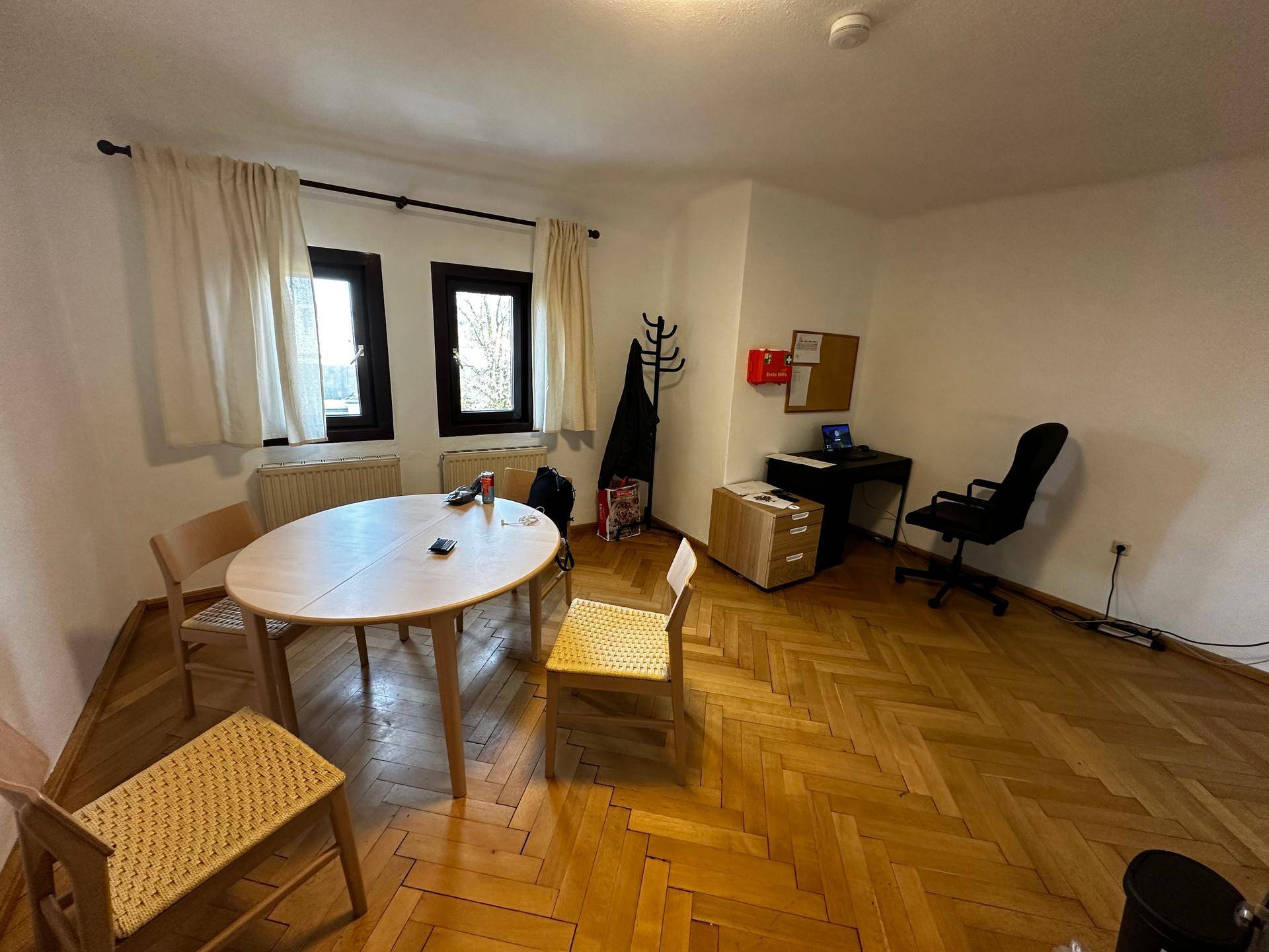 Esszimmer mit ovalem Holztisch, Stühlen und einem Arbeitsbereich am Fenster mit Parkettboden.