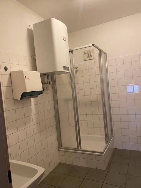 Kleines Badezimmer mit Duschkabine, weiß gefliesten Wänden und einem Warmwasserboiler an der Wand.