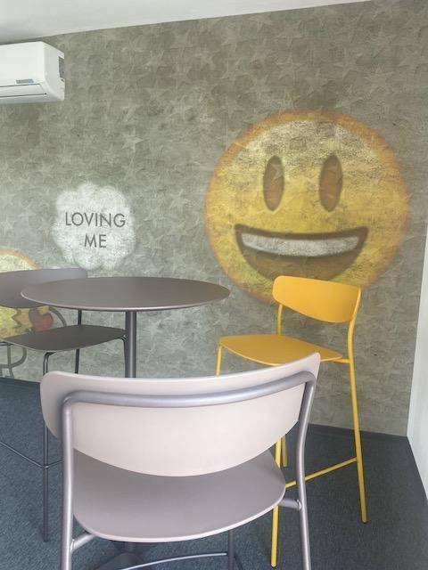 Heller Loungebereich mit gelben Stühlen, Tischen und einer Wanddekoration mit Smiley-Motiv und Klimaanlage.