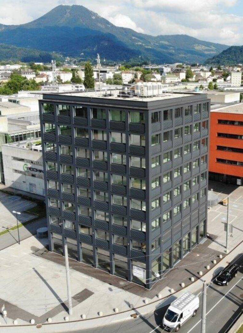 Luftaufnahme eines modernen Bürogebäudes mit Bergpanorama und städtischer Umgebung im Hintergrund.