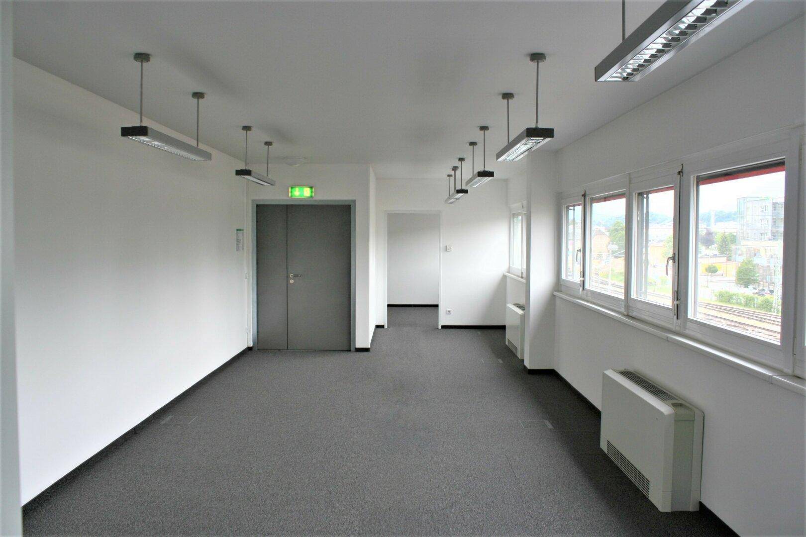 Leerer Büroraum mit grauem Teppichboden, großen Fenstern und Deckenbeleuchtung, bereit zur Einrichtung.