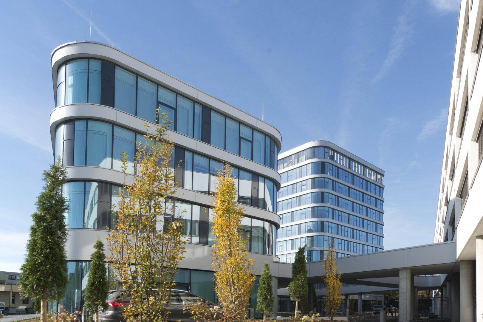 Moderne Bürogebäude mit geschwungenen Glasfassaden und umliegender Landschaftsgestaltung unter blauem Himmel.