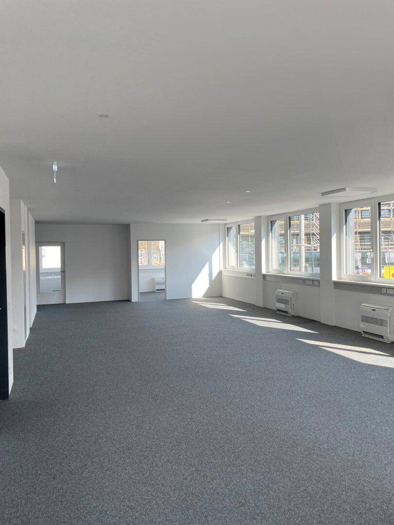 Großer, heller Büroraum mit grauem Teppichboden, vielen Fenstern und viel Tageslicht.