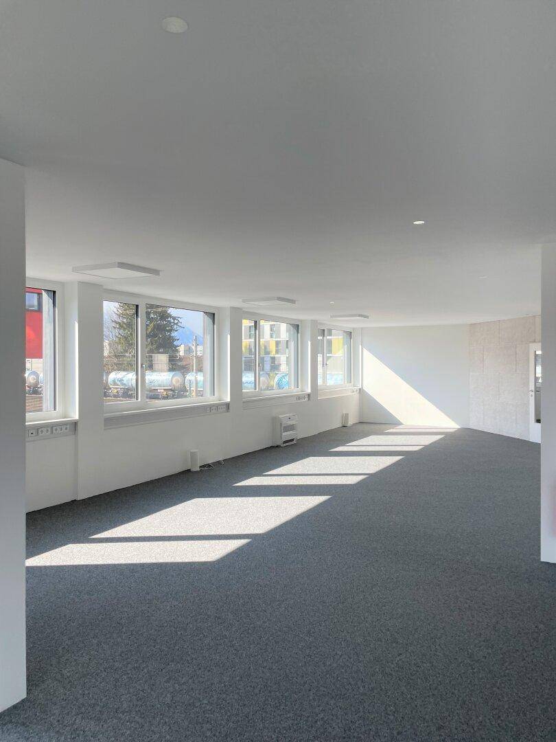 Heller Büroraum mit gebogener Wand, grauem Teppich und viel Tageslicht durch große Fenster.