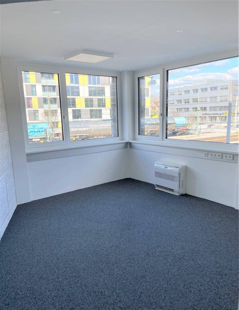 Heller Eckraum mit grauem Teppichboden, großen Fenstern und urbanem Ausblick auf Gebäude.