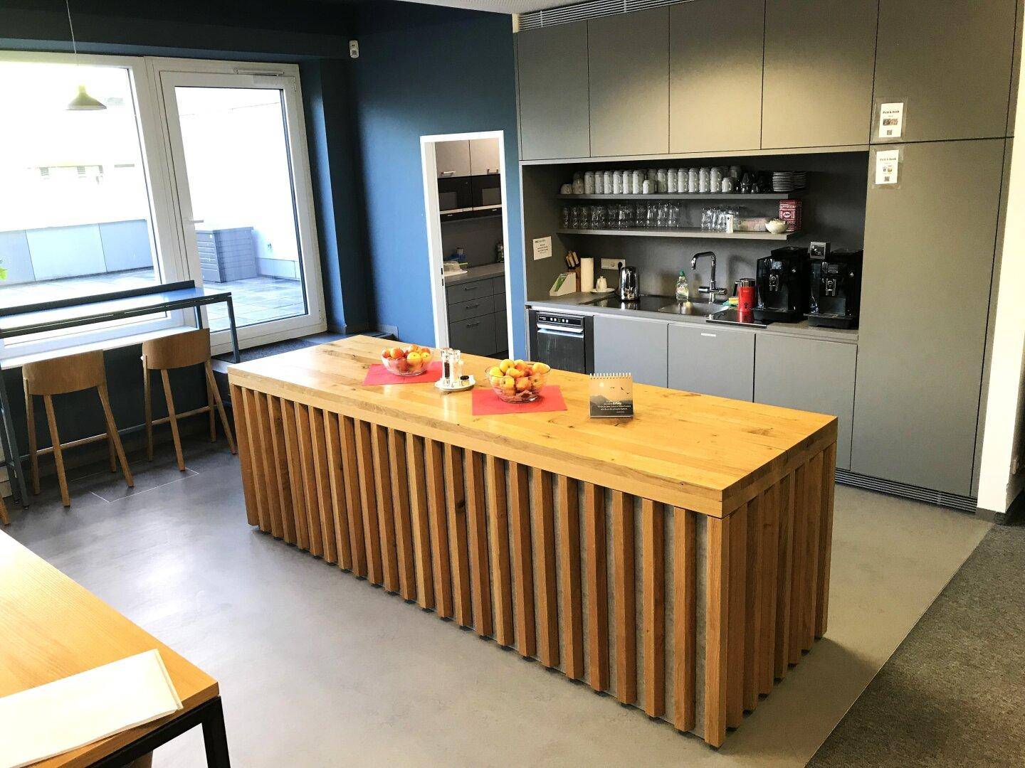 Moderne Teeküche mit Holztisch, grauen Schränken und Zugang zu einer Außenterrasse.