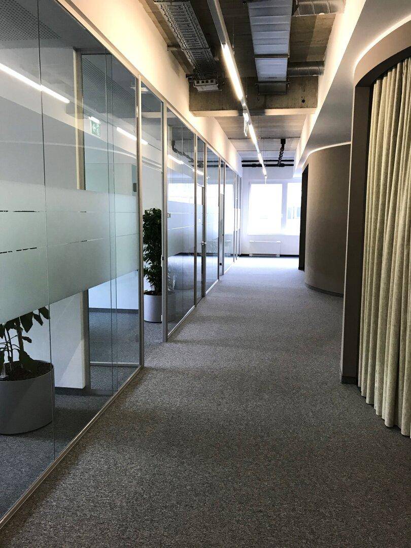 Langer Bürogang mit Glaswänden zu den Büros und Teppichboden, gut beleuchtet.