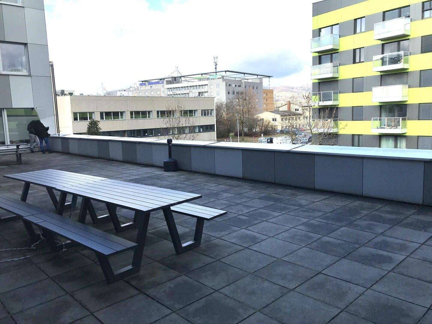 Große Außenterrasse mit Holzbänken und Tischen, umgeben von Gebäuden und Blick auf die Umgebung.
