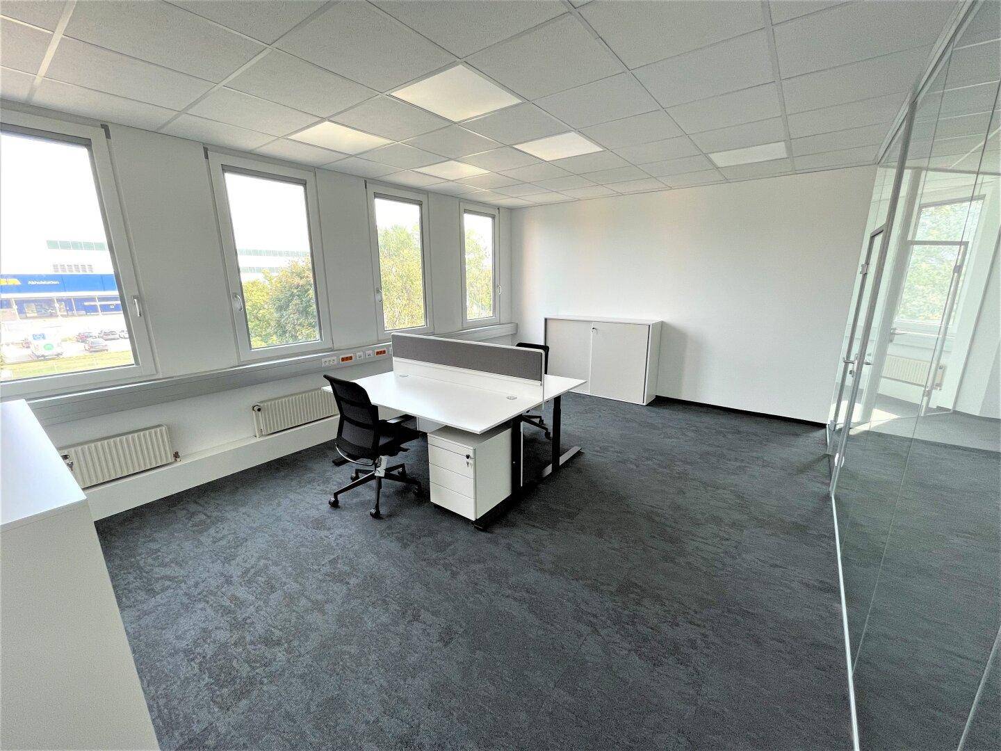 Einzelbüro mit Schreibtisch, Bürostuhl und großen Fenstern, die viel Tageslicht hereinlassen.