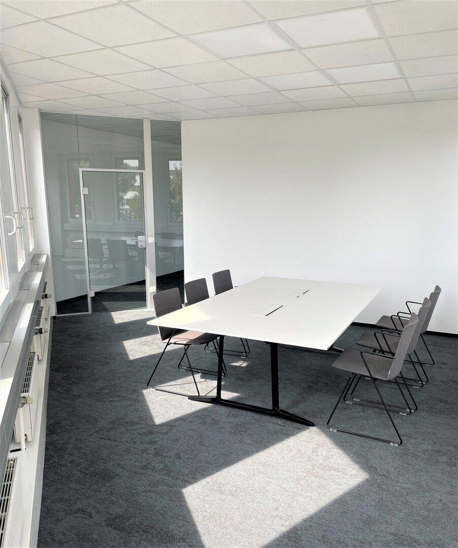 Moderner Besprechungsraum mit großem Tisch und Stühlen, umgeben von Glaswänden und großen Fenstern.
