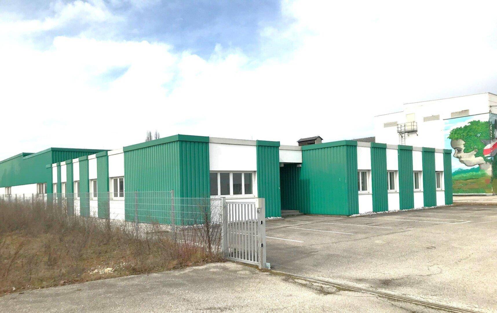Gebäude mit grün-weißer Fassade und Parkplatz davor, umgeben von einem Zaun. Im Hintergrund ist ein weiteres Gebäude mit einem großen Wandgemälde zu sehen.