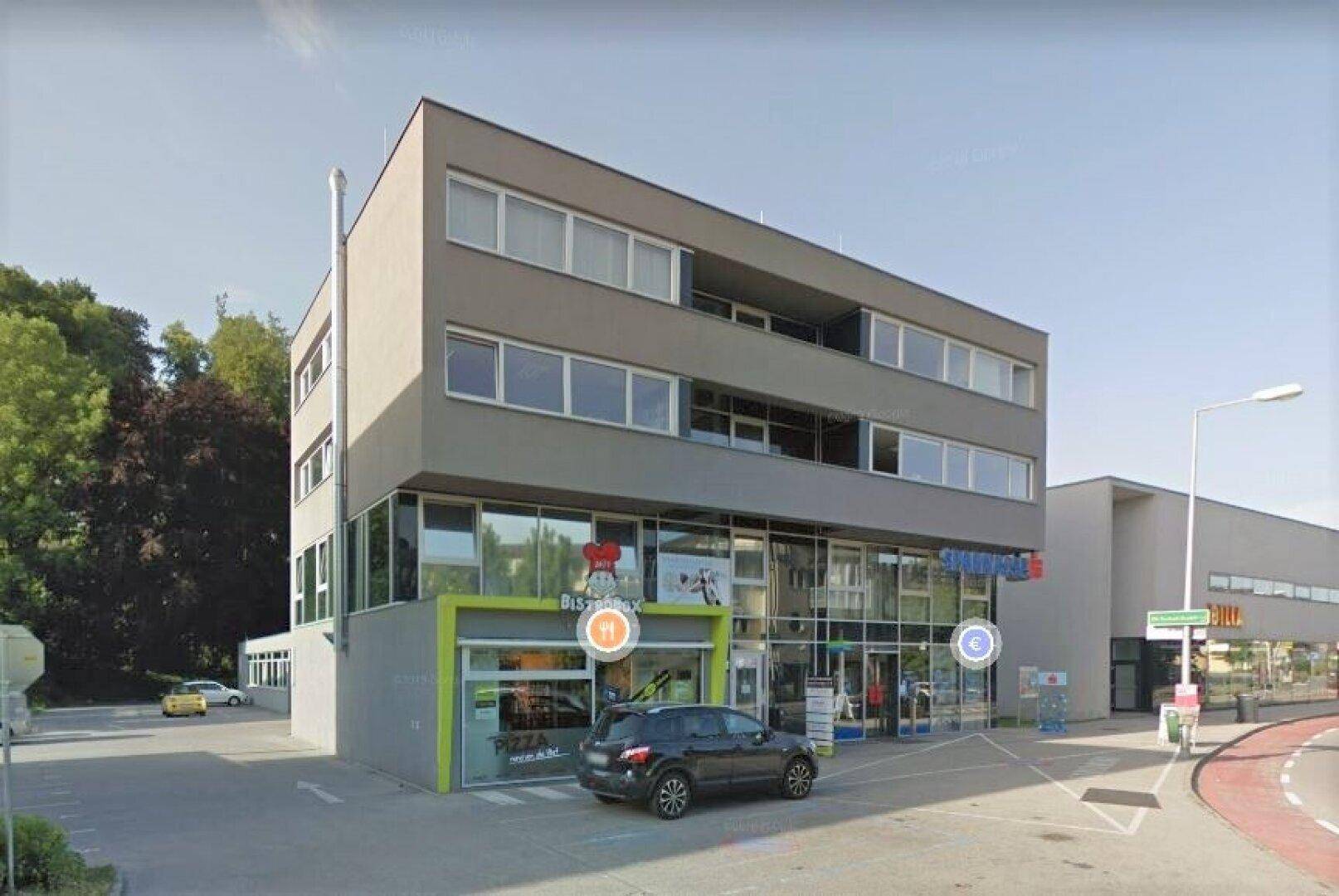 Ansicht eines modernen Gebäudes mit Büros und Geschäften im Erdgeschoss, umgeben von Grünflächen.