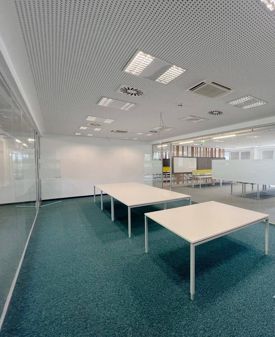 Heller Büroraum mit Teppichboden, großen Tischen und Glaswänden, ausgestattet mit moderner Deckenbeleuchtung.