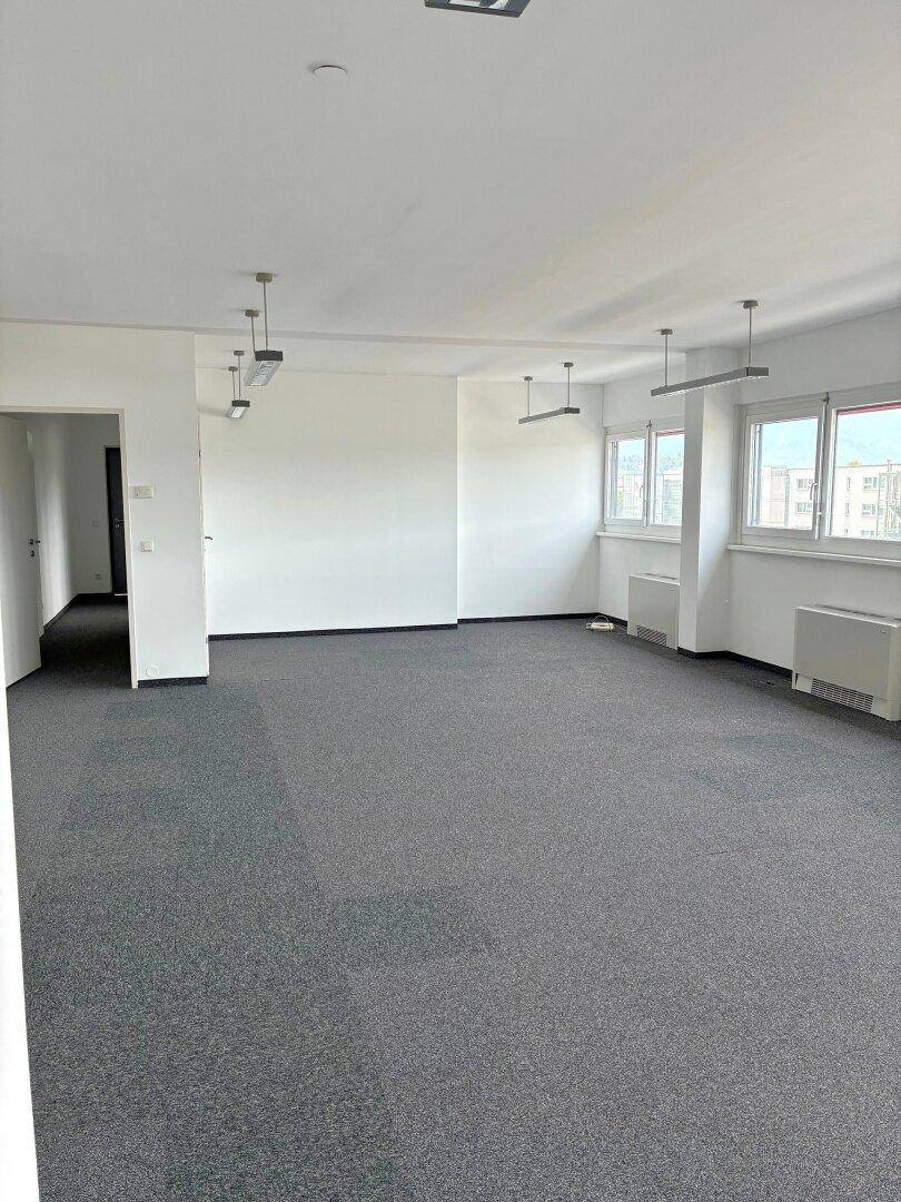 Geräumiges, leeres Büro mit Teppichboden, großen Fenstern und moderner Deckenbeleuchtung.