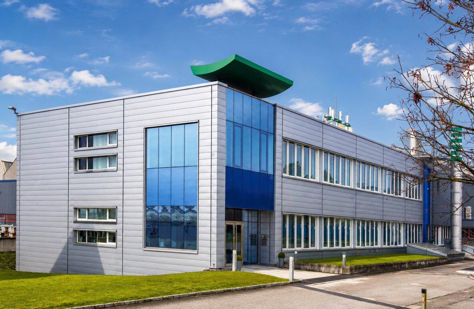 Modernes Bürogebäude mit Glasfassade, blauen Akzenten und gepflegter Grünfläche davor.
