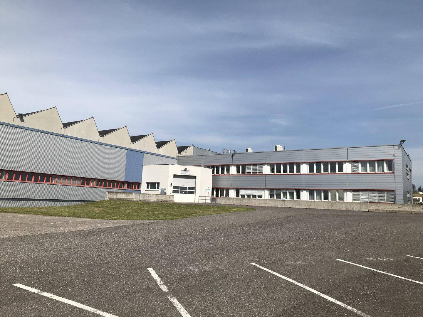 Industriekomplex mit großem Parkplatz und gezacktem Dach unter bewölktem Himmel.