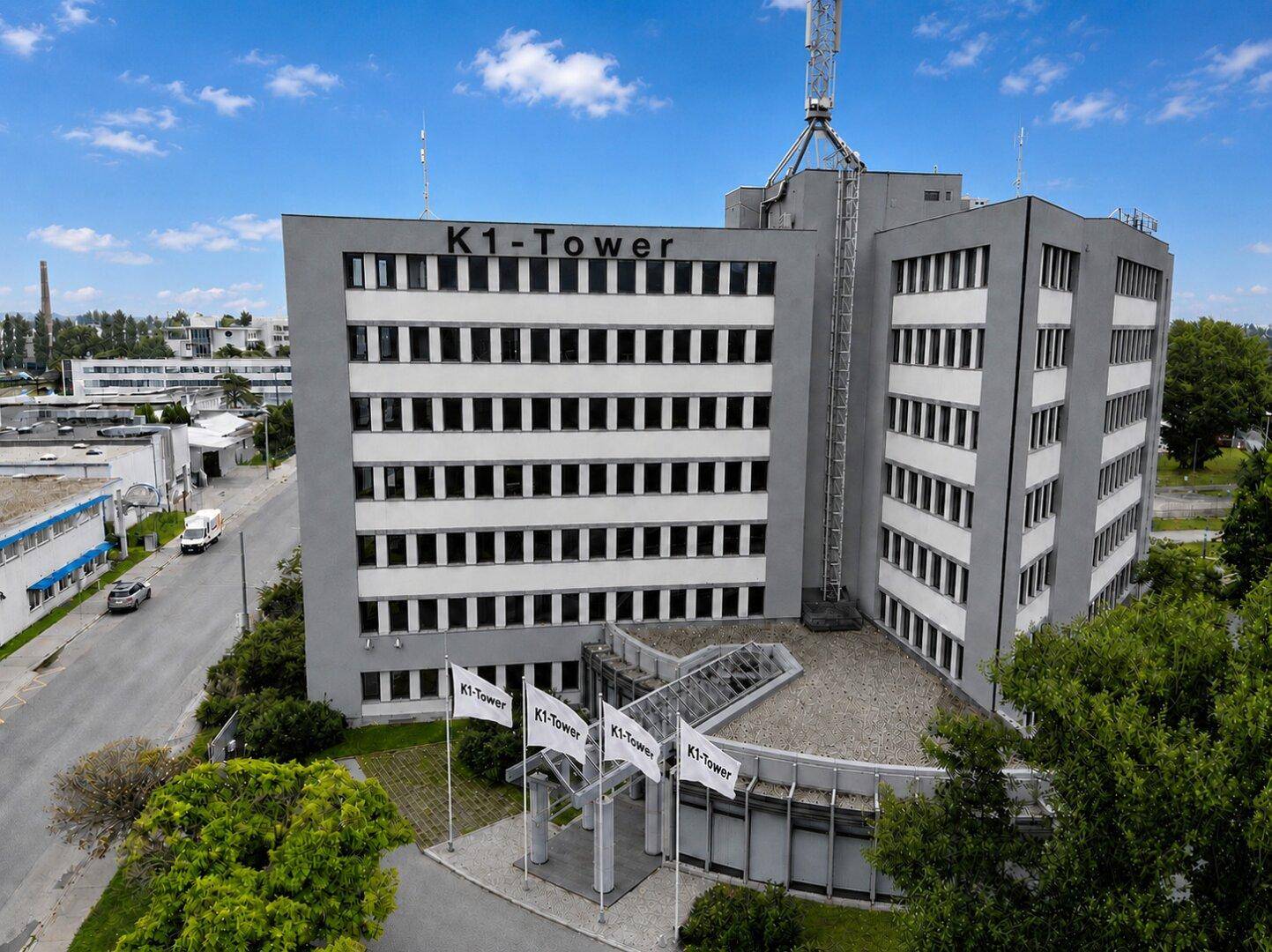 Blick auf den K1-Tower, ein modernes Bürogebäude mit grauer Fassade und vielen Fenstern, umgeben von Grünflächen und einer Zufahrtsstraße.