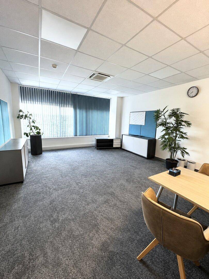 Helles und gepflegtes Büro mit grauem Teppichboden, vertikalen Jalousien und modernen Möbeln in einem Gewerbeobjekt.