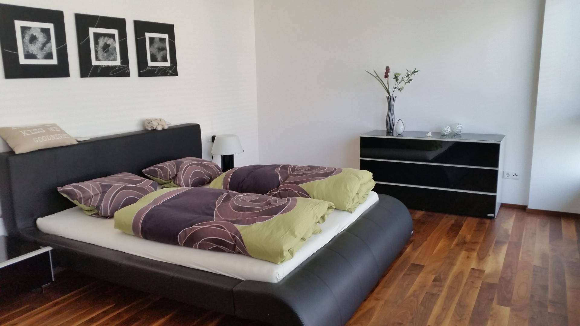 Schlafzimmer mit großem Doppelbett, Holzboden und moderner Kommode in Schwarz.