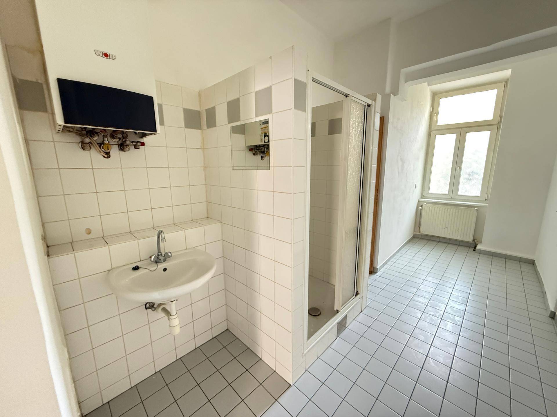 Badezimmer mit Waschbecken, Duschkabine und weißen Fliesen, gut beleuchtet durch ein Fenster.