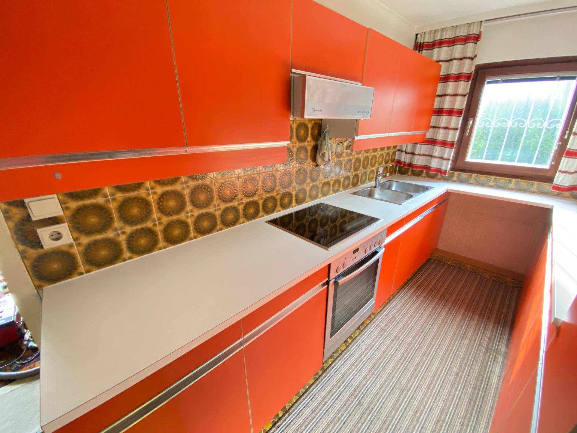 Auffällige Küche mit leuchtend orangefarbenen Schränken, gemusterten Fliesen und einem modernen Induktionskochfeld.