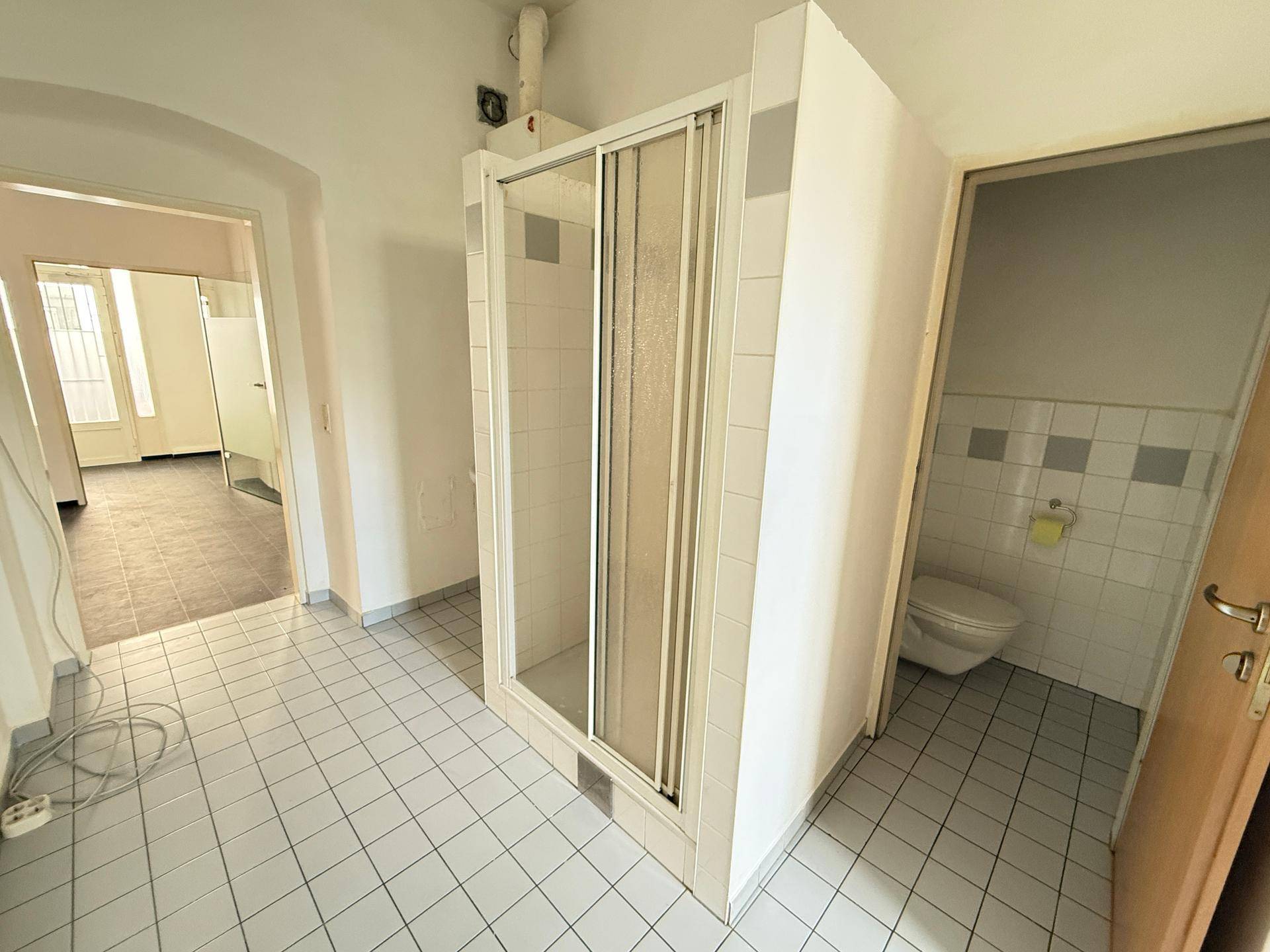 Badezimmer mit Duschkabine, Toilette und weißen Fliesen, angrenzend an weitere Räume.