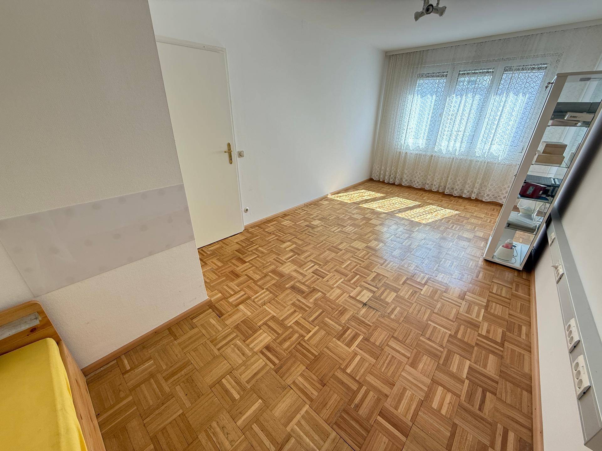 Schlafzimmer mit Parkettboden, Fenster mit hellen Vorhängen und einem großen Standspiegel.