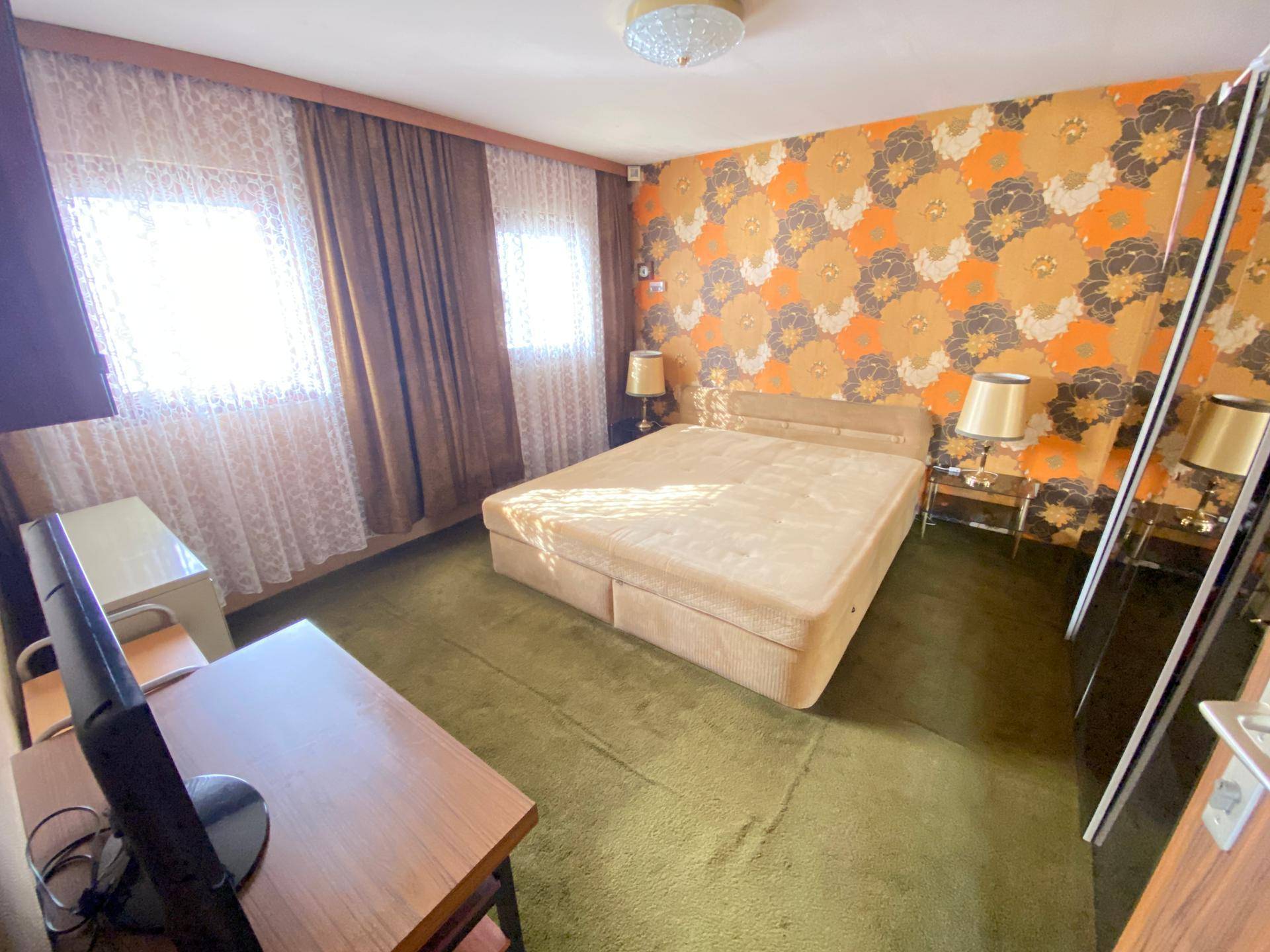Geräumiges Schlafzimmer mit großem Bett, grünem Teppichboden und auffälliger floraler Tapete, hell beleuchtet.