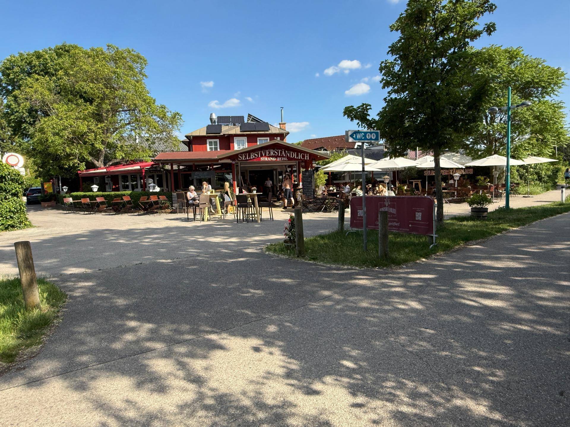 Außenbereich eines Restaurants oder Cafés mit Tischen und Sonnenschirmen, umgeben von Bäumen.