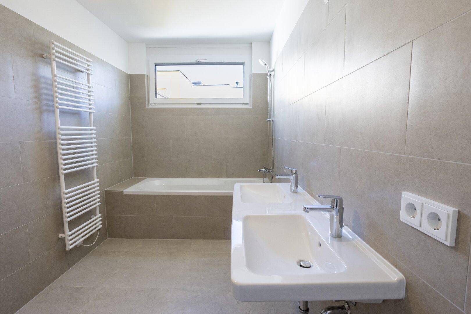 Geräumiges Badezimmer mit Badewanne, Doppelwaschbecken und beheiztem Handtuchhalter, hell gefliest.