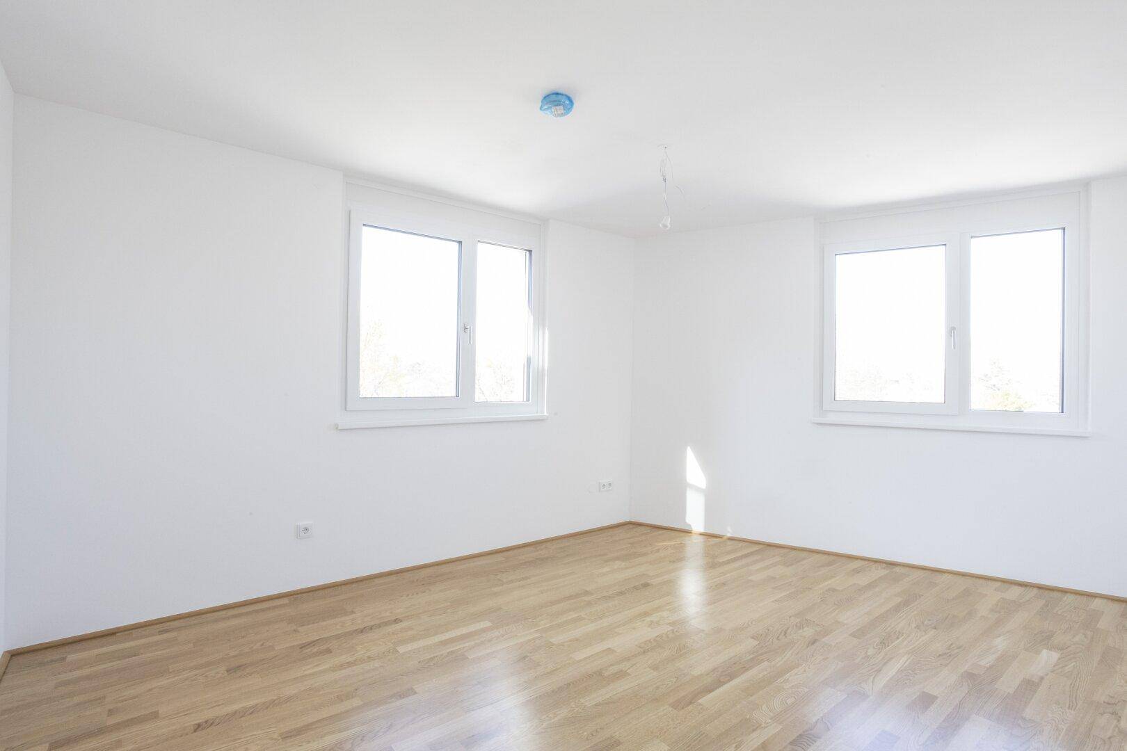 Lichtdurchflutetes Schlafzimmer mit Parkettboden und zwei Fenstern, schafft eine helle Wohnatmosphäre.