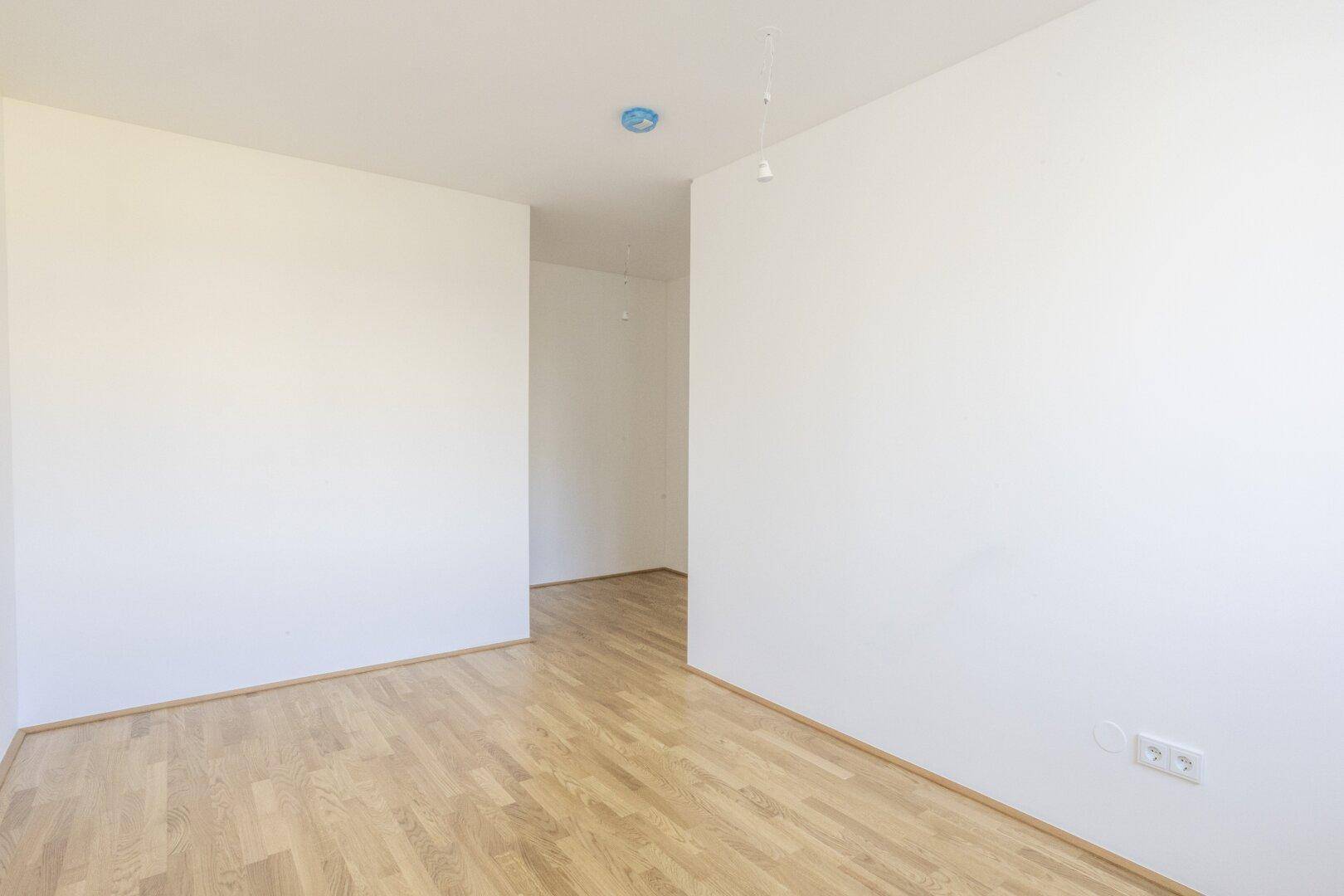 Heller Raum mit Holzboden und weiß gestrichenen Wänden, offen zu einem weiteren Bereich.