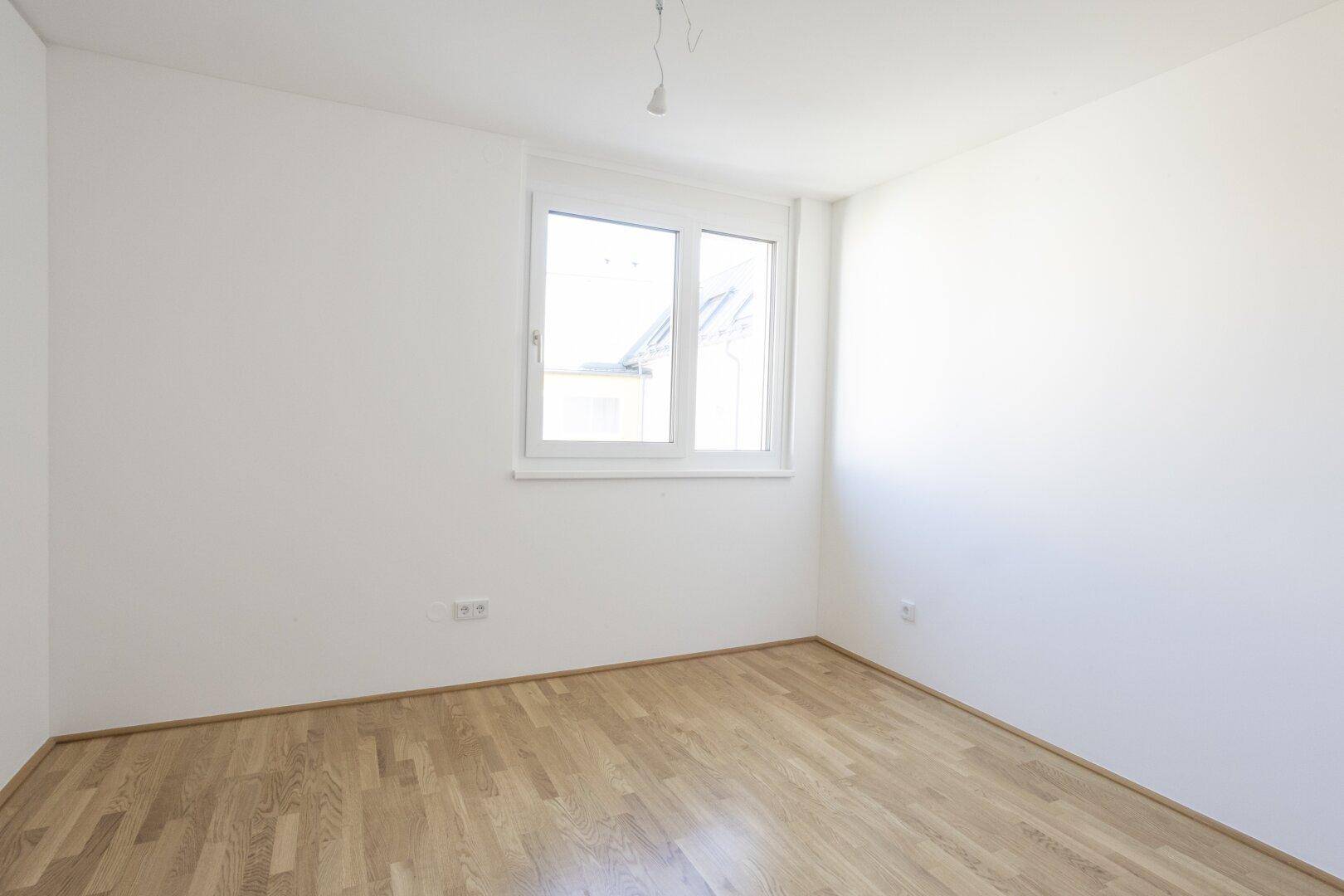 Helles Schlafzimmer mit Holzboden und einem großen Fenster, das viel Tageslicht hereinlässt.