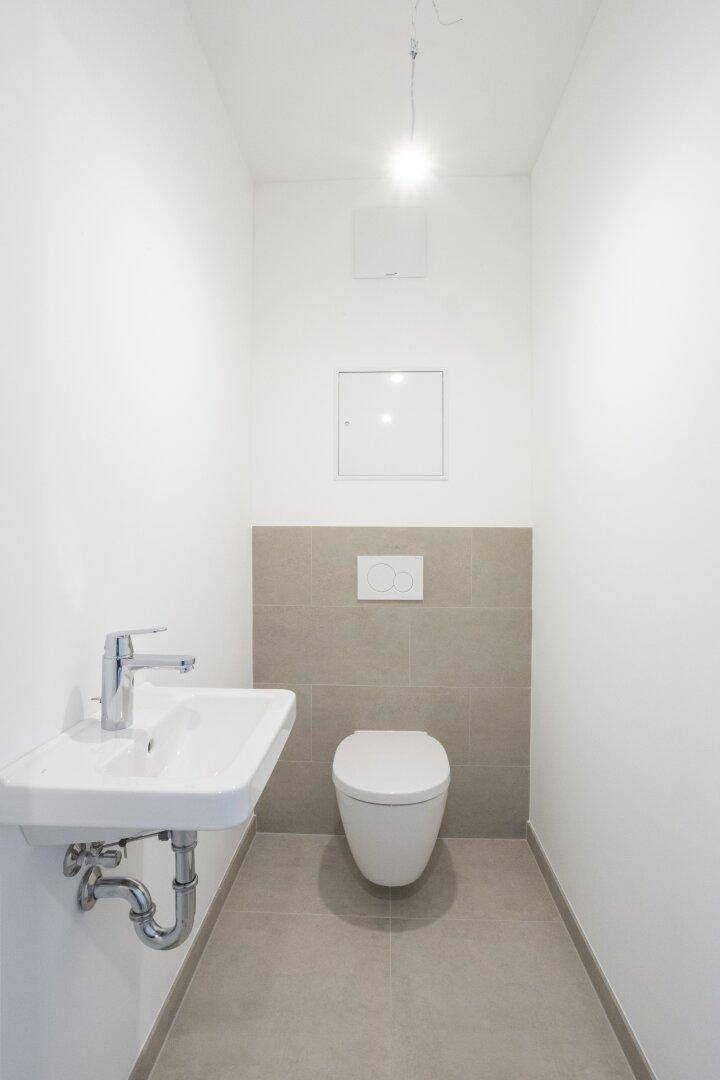 Modernes Gäste-WC mit Wand-WC, kleinem Waschbecken und hellgrauen Fliesen an der Wand.