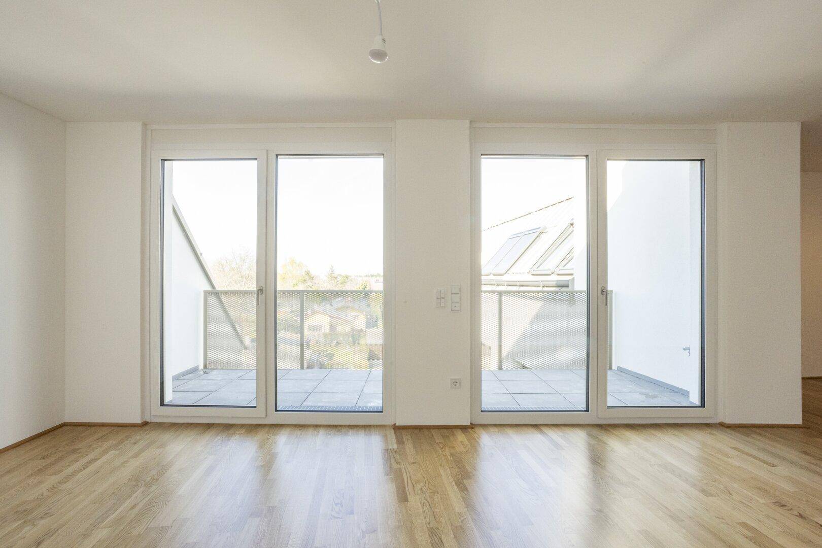Helles Zimmer mit Holzboden und breiter Fensterfront, die auf eine Terrasse blickt.