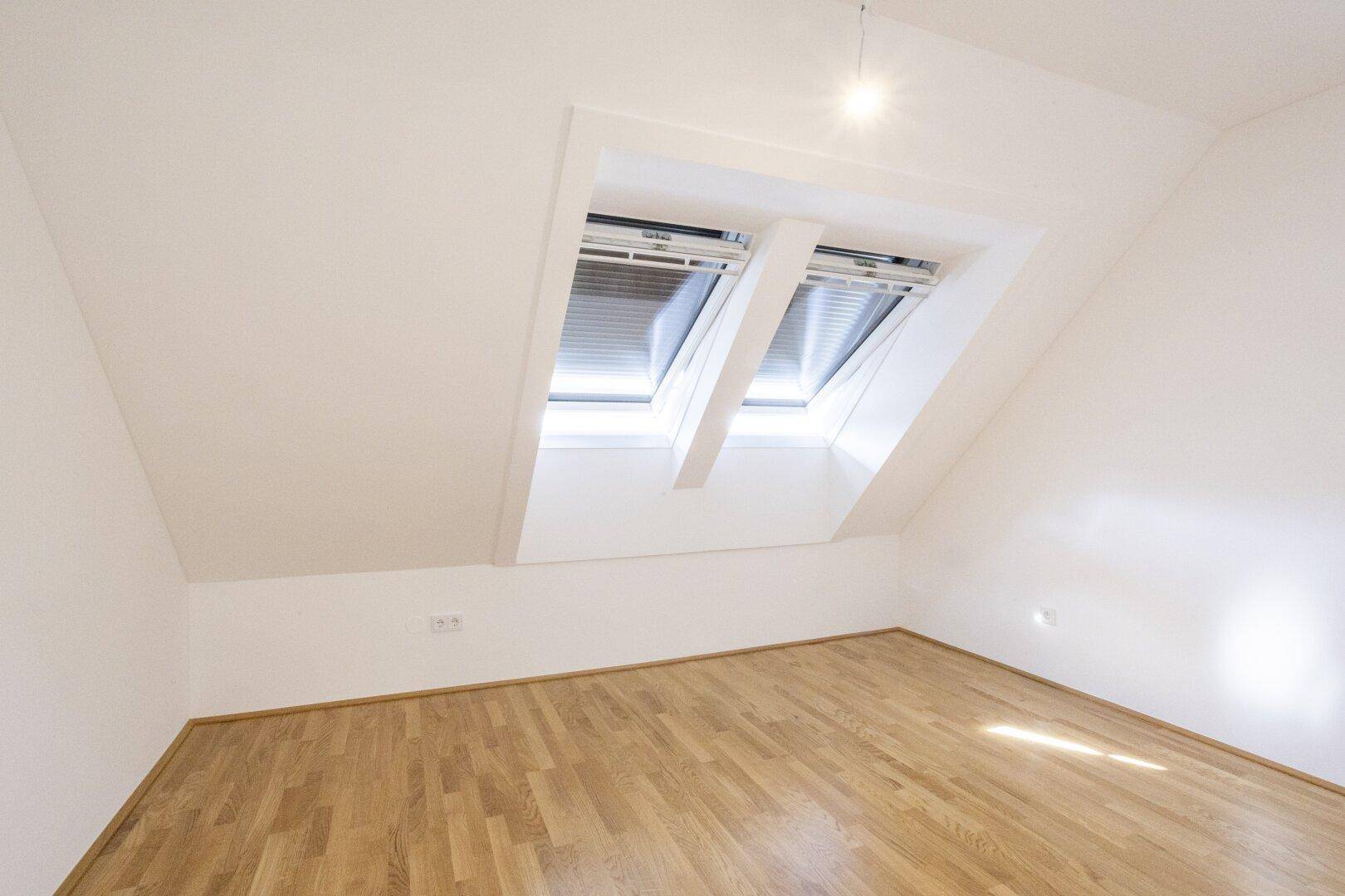 Helles Dachzimmer mit Holzboden und zwei Dachfenstern, die den Raum mit Licht füllen.