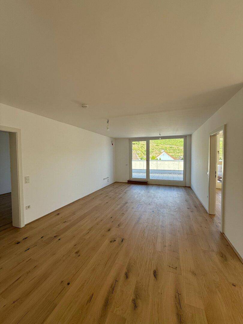 Geräumiges Wohnzimmer mit hellem Holzfußboden, großen Fenstern und Zugang zur Terrasse mit Blick ins Grüne.