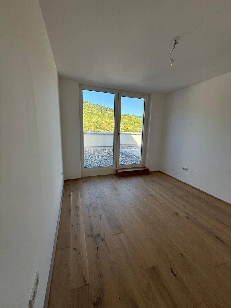 Leeres Schlafzimmer mit Holzboden, großem Fenster und Zugang zu einer Terrasse mit Weinbergblick.