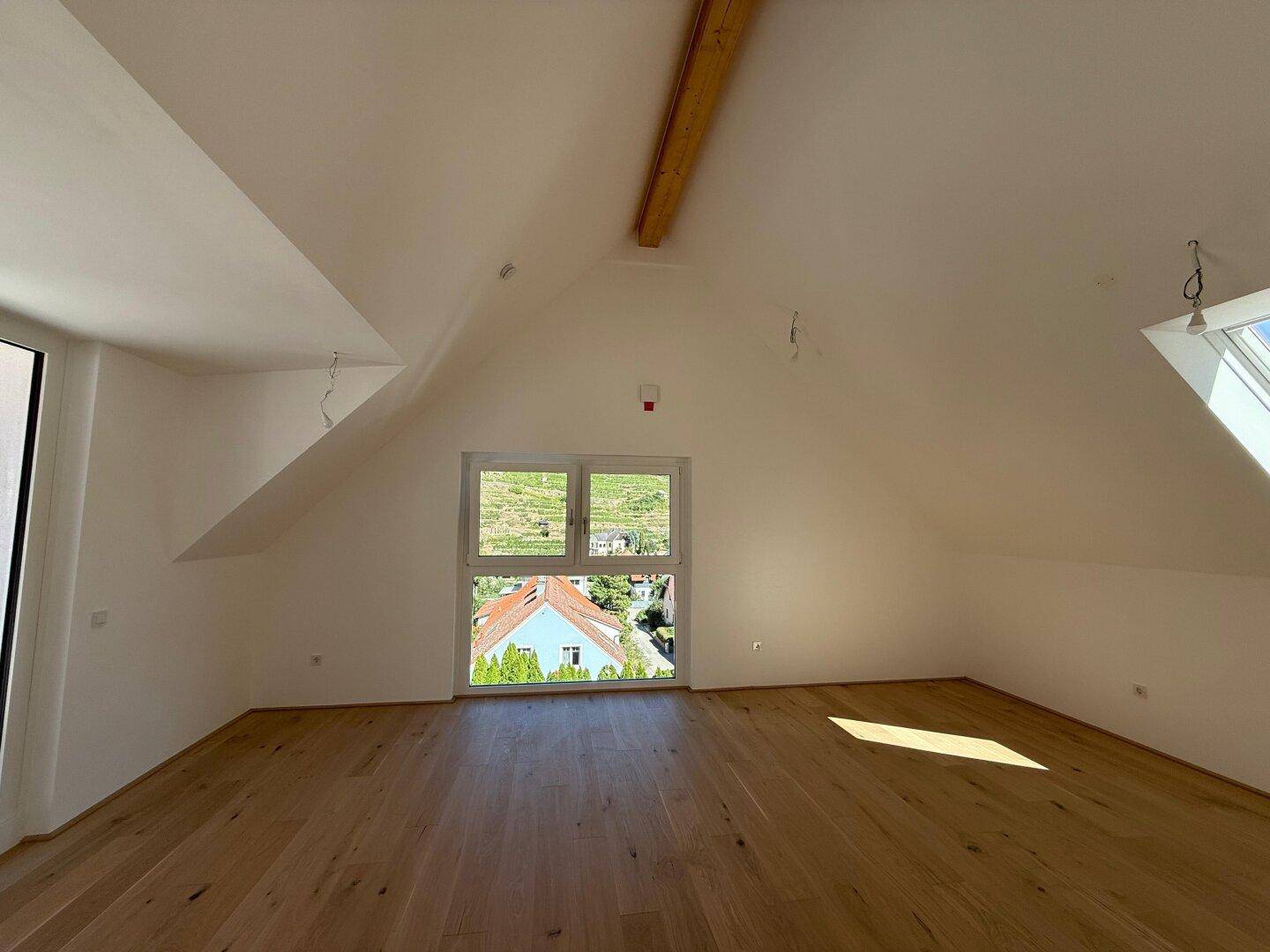 Geräumiger Dachgeschossraum mit schrägen Wänden, Holzbalken und weitem Ausblick ins Grüne.