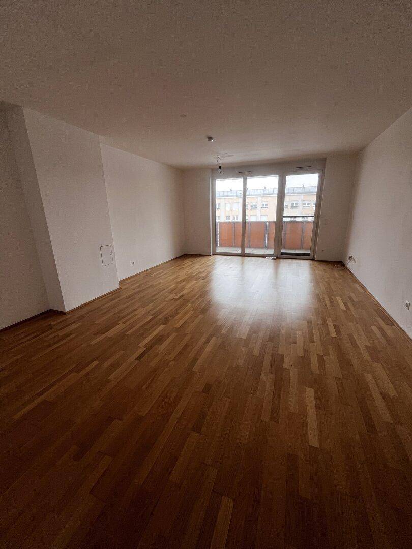 Geräumiges, leeres Wohnzimmer mit hellem Parkettboden und Zugang zu einem Balkon.
