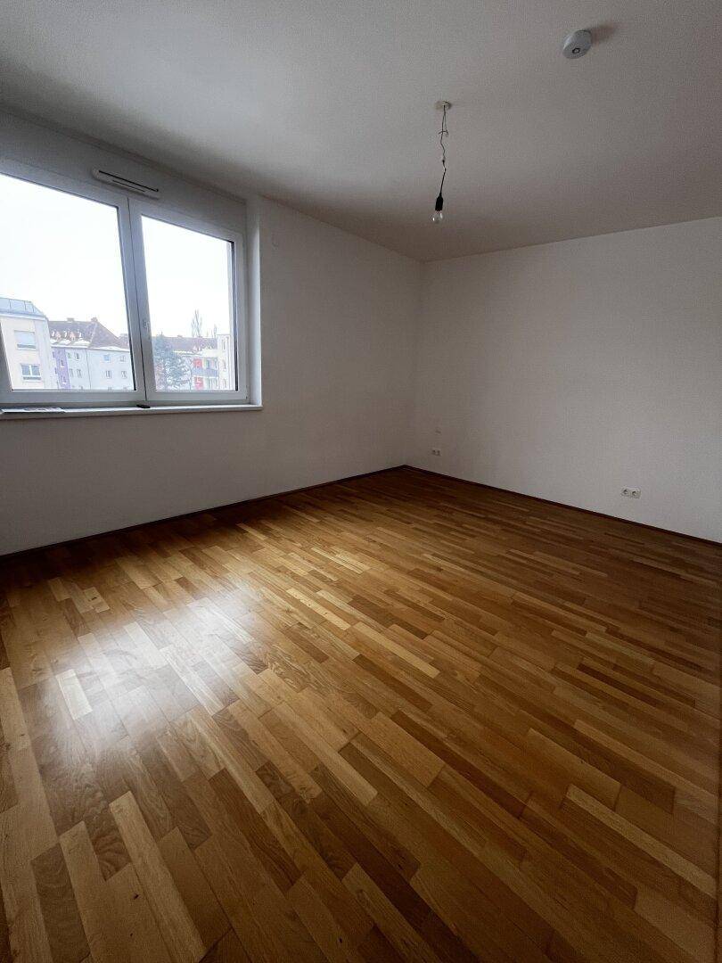 Leeres Schlafzimmer mit Parkettboden und einem großen Fenster, das viel Licht hereinlässt.
